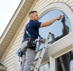 Professional window cleaner on a ladder using a squeegee to clean an exterior arched window, San Antonio, TX.